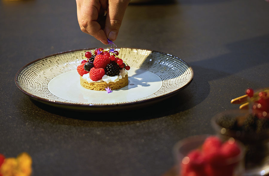 Une main experte pose une pétale de plus sur une tarte aux fruits rouges raffinée posée dans une assiette blanche aux motifs marrons