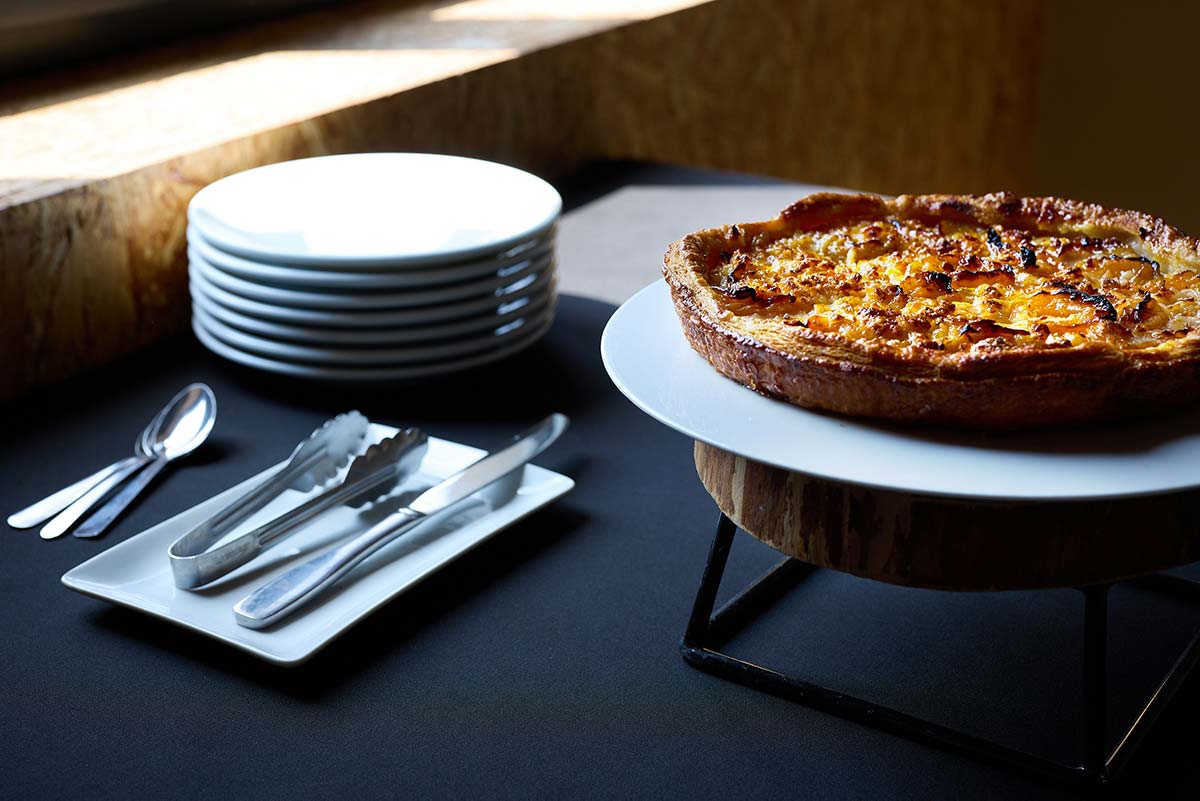 Une belle tarte présentée sur un support surelevé et des couverts et assiettes posés à côté pour se servir sur le buffet